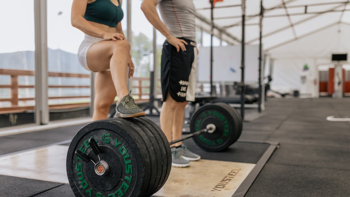 Two adults in a gym setting with weightlifting equipment and sportswear, focused on strength training.